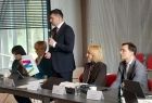 Marszałek Łukasz Smółka stoi przy stole i trzyma mikrofon. Obok siedzą inni ludzie.