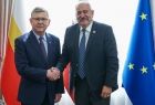 Wicemarszałek Witold Kozłowski stoi z mężczyzną i podaje mu rękę. Za nimi widoczne flagi.