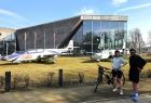 widok na gmach główny Muzeum Lotnictwa Polskiego, przed gmachem stoją samoloty, na pierwszym planie kobieta i mężczyzna w kaskach rowerowych oraz rower