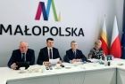 Zdjęcie przedstawia czterech członków Zarządu Województwa Małopolskiego, siedzących przy stole konferencyjnym. Za nimi, na ścianie, widnieje duże logo Województwa Małopolskiego.