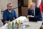 Wicemarszałek Ryszard Pagacz siedzi przy stole z mężczyzną. Za nim widoczne są flagi.