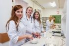 Zdjęcie przedstawia pięć kobiet w białych fartuchach, stojących przy długim stole, w tle wisi zielona tablica kredowa.