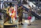 Sztandar Surge Polonia w gablocie trzymany przez manekina. Przy gablocie stoją mężczyzna i kobieta w mundurach wojskowych i rozmawiają ze sobą