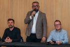 Dwie osoby siedzące przy stole konferencyjnym, jedna stoi i mówi do mikrofonu.