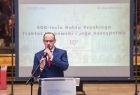 Zdjęcie osoby przy mikrofonie, za nim slajd wyświetlany na ekranie z nazwą konferencji.
