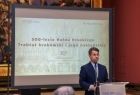 Zdjęcie osoby przy mikrofonie, za nim slajd wyświetlany na ekranie z nazwą konferencji.