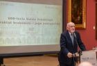 Zdjęcie osoby przy mikrofonie, za nim slajd wyświetlany na ekranie z nazwą konferencji.