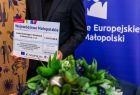 Symboliczny czek prezentowany na tle baneru Funduszy Europejskich