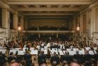 Zdjęcie przedstawia muzyków Filharmonii Krakowskiej podczas koncertu na estradzie koncertowej, usytuowanych tyłem do fotografującego, a przodem do widowni.