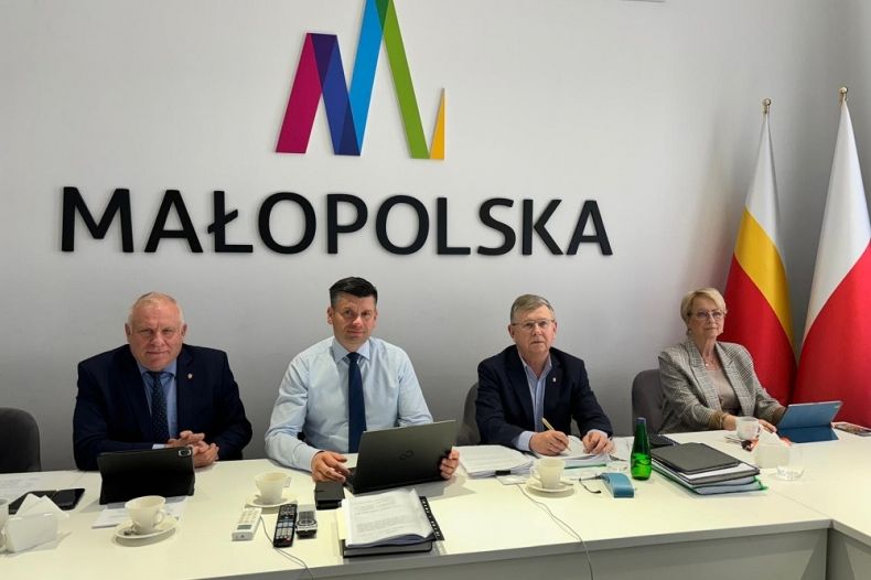 Zdjęcie członków Zarządu Województwa Małopolskiego, siedzących przy stole konferencyjnym, za ich plecami duże logo Województwa Małopolskiego.