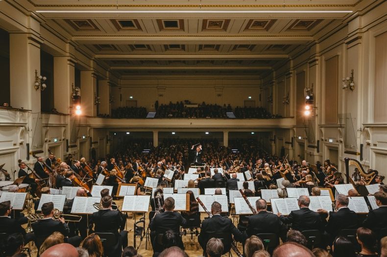 Zdjęcie przedstawia muzyków Filharmonii Krakowskiej podczas koncertu na estradzie koncertowej, usytuowanych tyłem do fotografującego, a przodem do widowni.