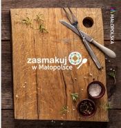 Na ciemnej desce drewnianej ułożona deska z jasnego drewna, na której rozsypana jest sól i gałązki rozmarynu. W prawym górnym rogu nóż i widelec. W prawym dolnym rogu dwie ciemne miseczki z przyprawami. Na środku biały napis Zasmakuj w Małopolsce.