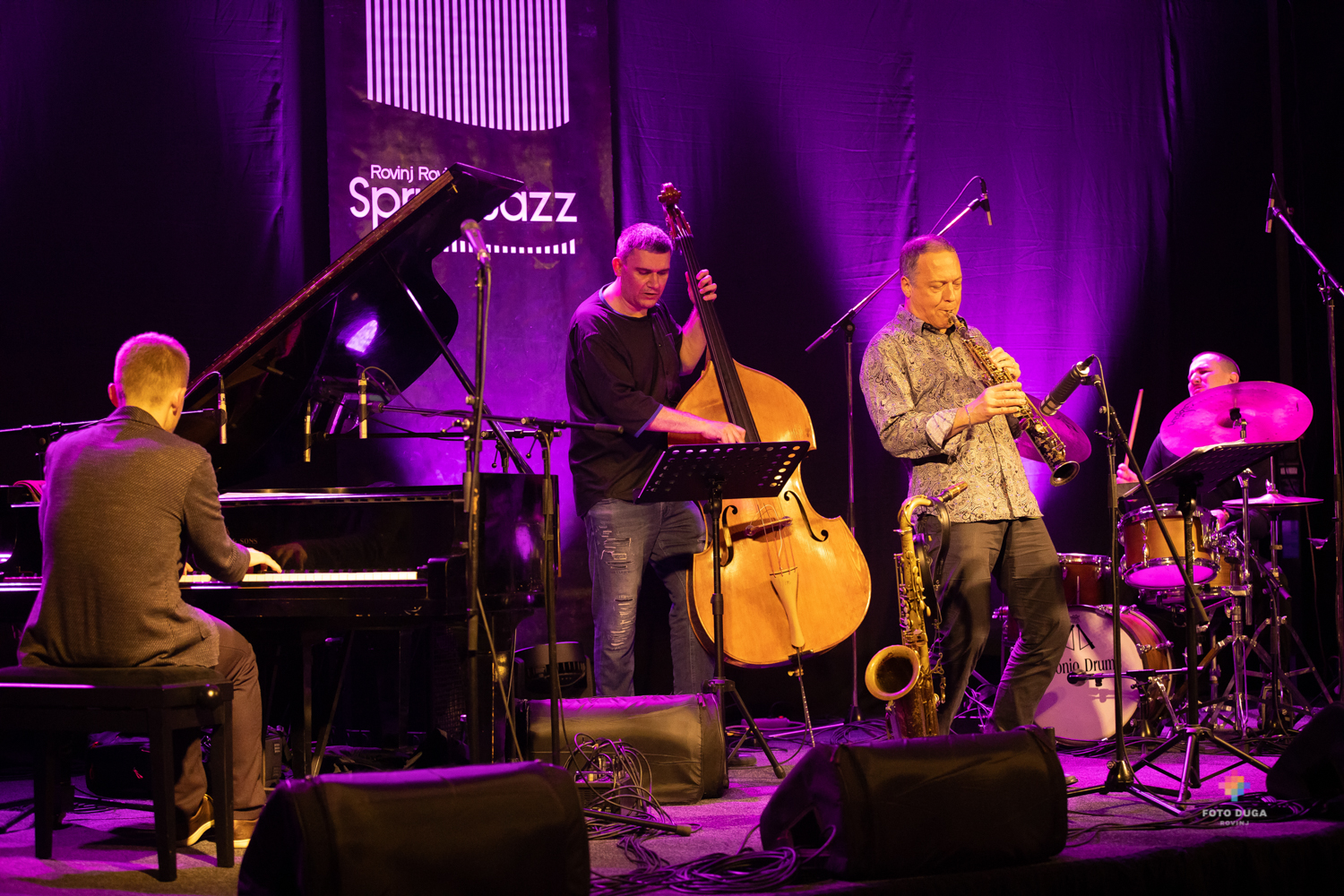 Na zdjęciu widać czterech muzyków grających koncert na scenie. Pianista siedzący przy pianinie, perkusista uderzający w talerz perkusji, saksofonista stojący na środku sceny oraz mężczyzna grający na kontrabasie, stojący za saksofonistą. Wszyscy bardzo wczuwają się w swoją grę. Inne ujęcie tej samej sceny jak na poprzednim zdjęciu. Tym razem na głównym planie widać tyłem siedzącego pianistę.