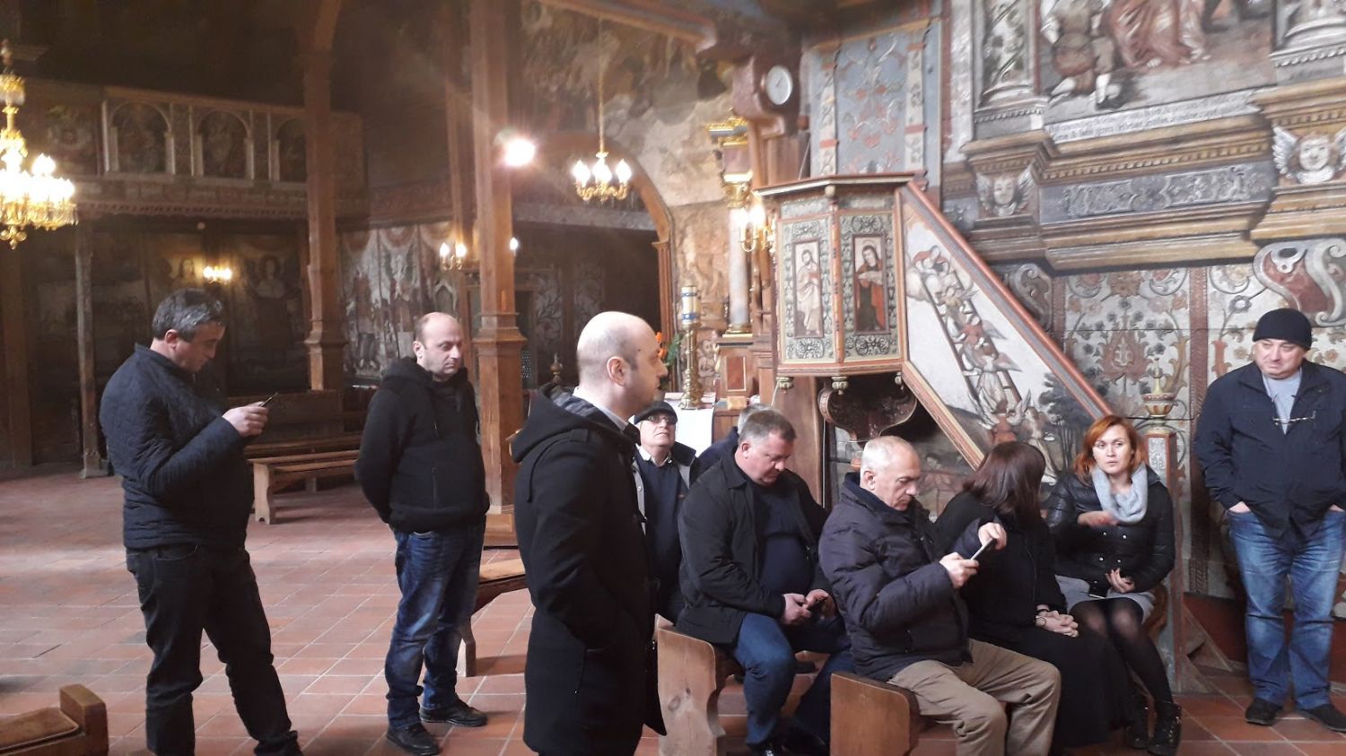 Na zdjęciu widać grupę osób w zabytkowym kościele. Część osób stoi i przygląda się freskom na ścianach, inne siedzą w ławach kościelnych.