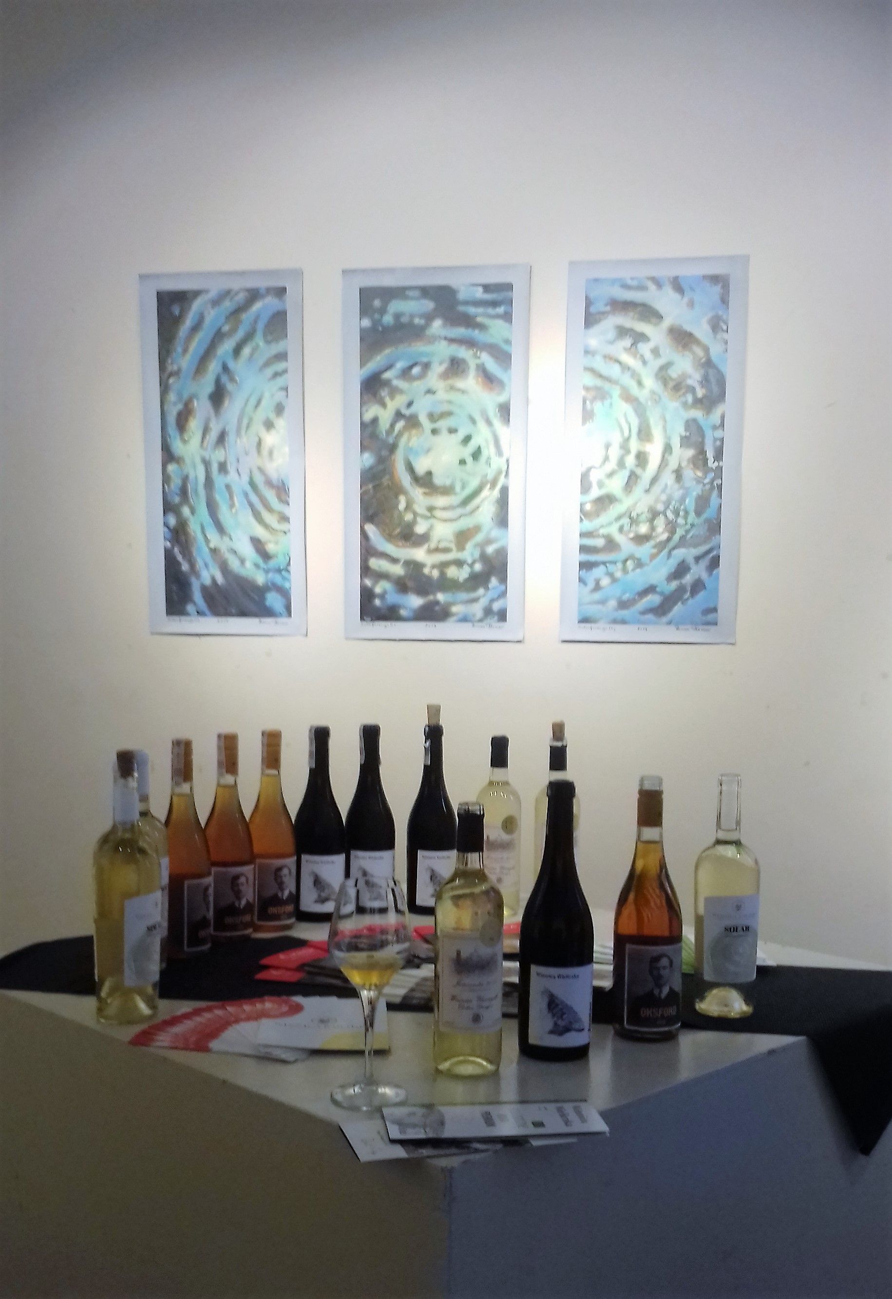 Na zdjęciu zbliżenie trzech obrazó na ścianie, Pod ścianą stoją w rzędzie butelki z polskimi winami.