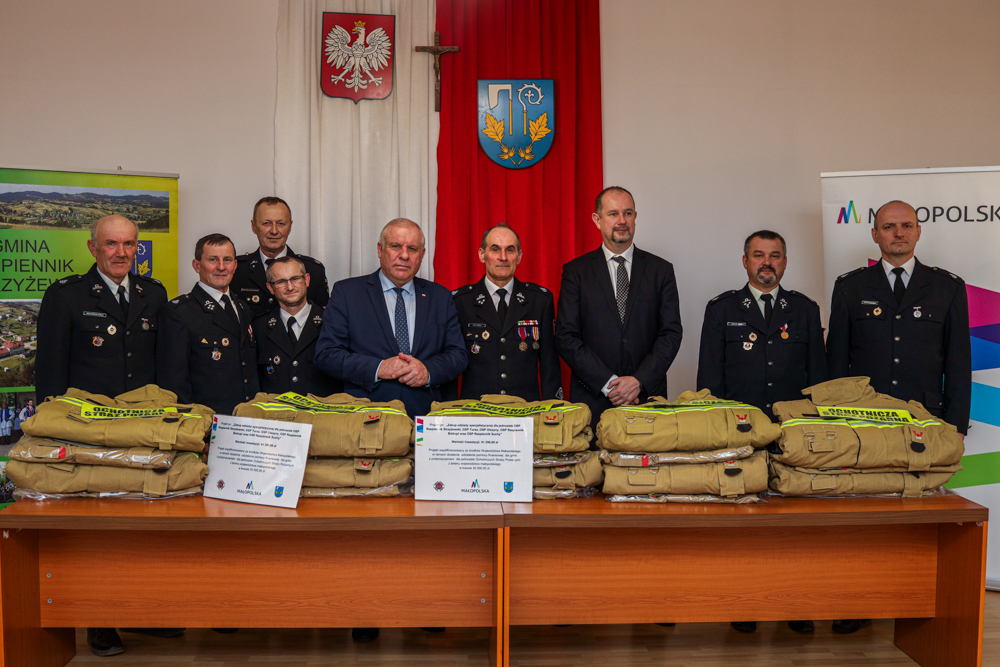 Uczestnicy wydarzenia stoją w sali. Na stołach widoczne umndury.