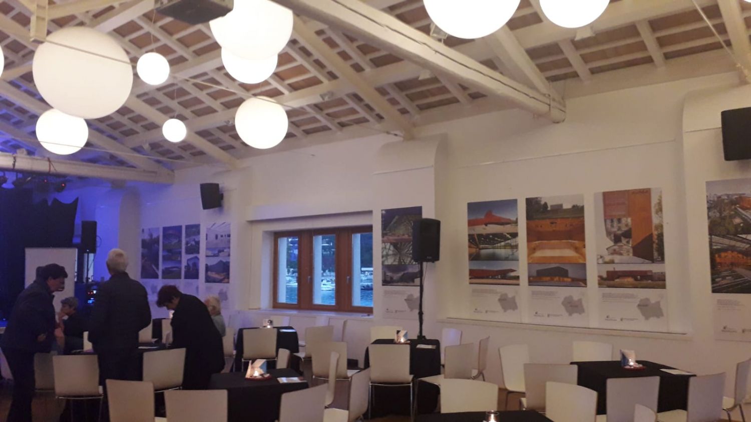 Na zdjęciu widać salę, na ścianach wiszą zdjęcia przedstawiające nowoczesne budynki z Małopolski, w sali znajdują sie stoliki z białymi krzesłami jak w restauracji, kilka osób stoi w rogu sali i dyskutuje.