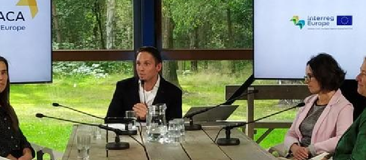 Grupa uczestników konferencji ITHACA siedzących przy stole i prowadzących rozmowę. W centrum zdjęcia mężczyzna moderujący dyskusję z trzema kobietami siedzącymi przy stole. W tle zieleń parkowa otaczająca ośrdek konferencyjny, położony niedaleko Eindhoven w Holandii.