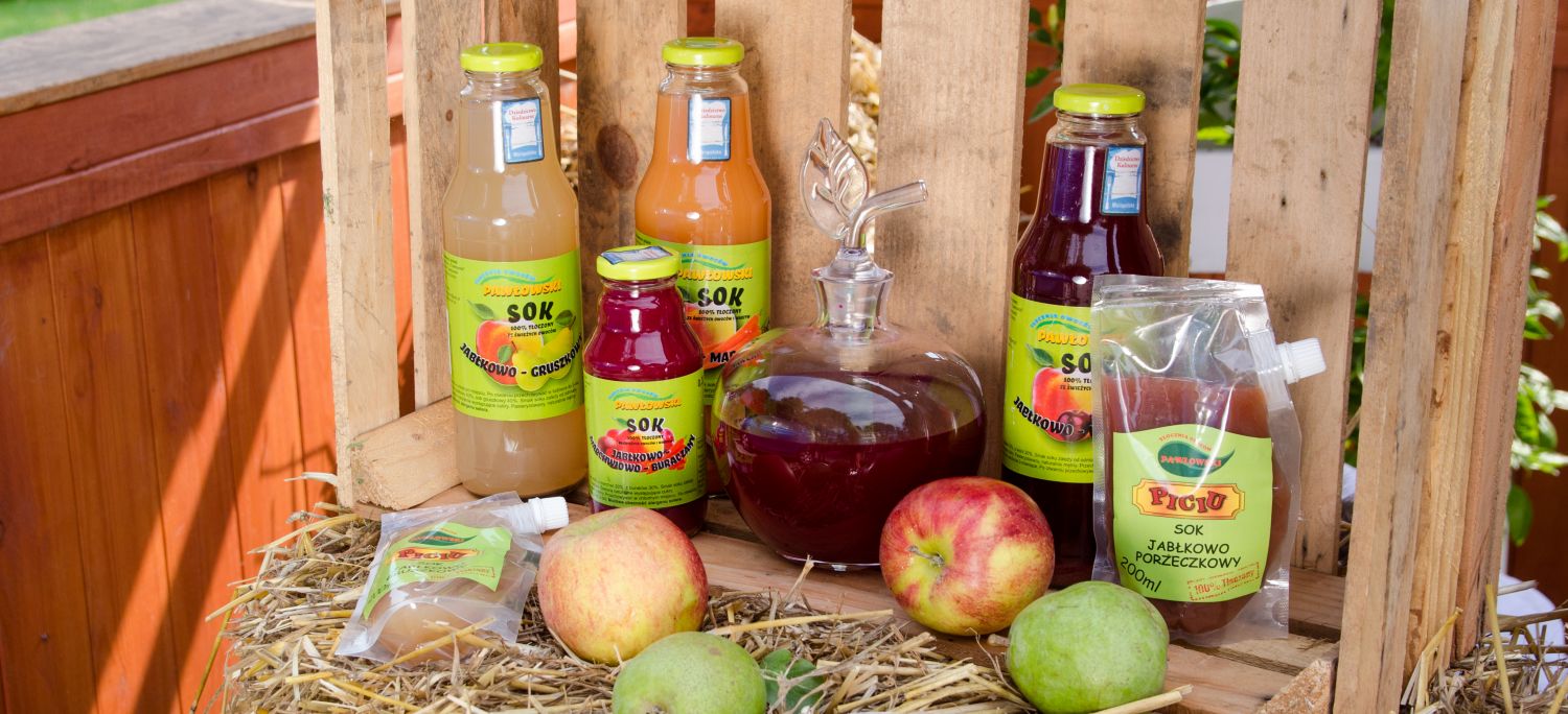 Na tle drewnianej paczki stoja butelki z różnymi sokami, Obok również leży kilka jabłek. Wszystko ustawione na słomie.