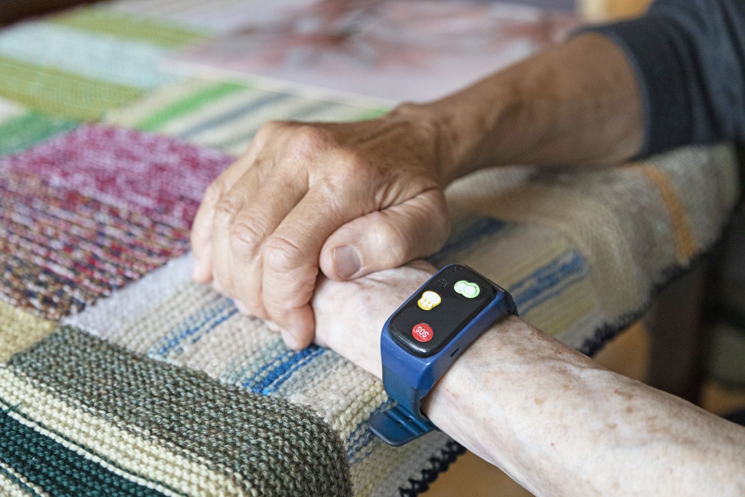 Dwie uściśnięte dlonie a na jeden z nich założona jest opaska bezpieczeństwa z widocznimi przyciskami do Centrum Teleopieki i alarmowym SOS.