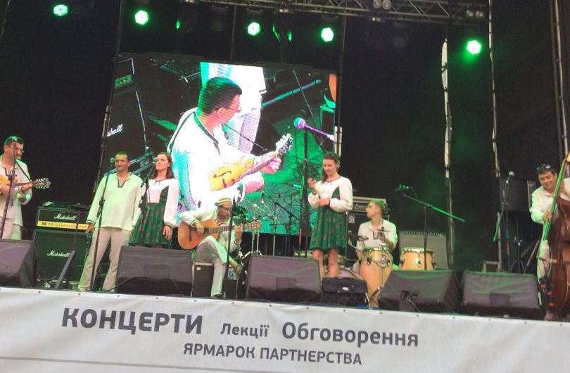 Kolorowe zdjęcie przedstawiające członków zespołu grających na instrumentach i śpiewających na scenie