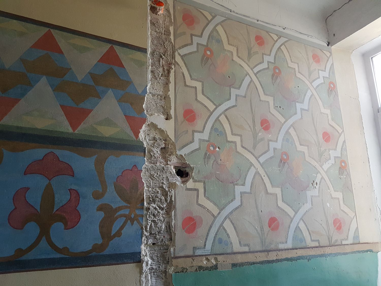 Na zdjęciu widać fragment ściany, na której widać odkryte podczas konserwacji fragmenty malowideł(szlaczki, figury różnej wielkości i koloru) i fragment zniszszonej ściany