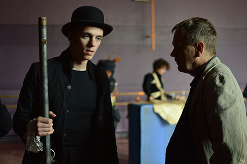 Na zdjęciu widać dwóch mężczyzn rozmawiających ze sobą, jeden z nich to aktor ubrany na czarno, w czarnym kapeluszu. 